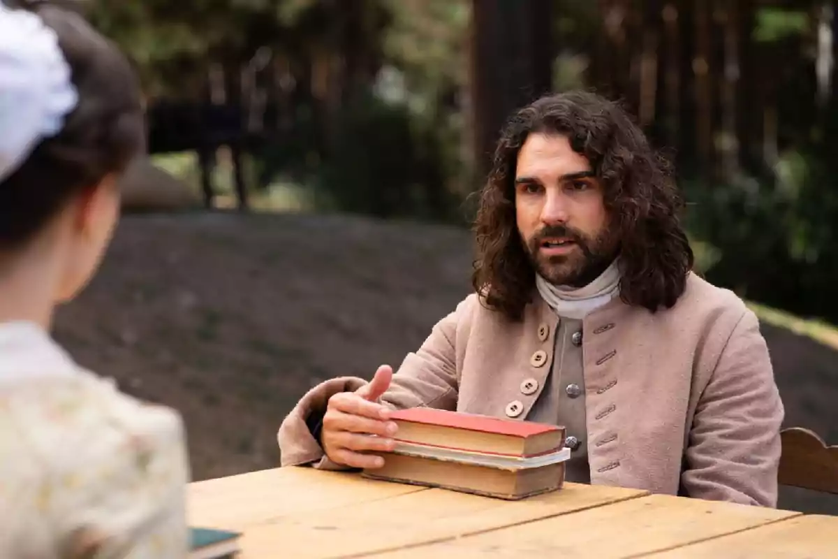 Sol con cabello largo y barba, vestido con ropa de época, está sentado al aire libre frente a una mesa de madera, sosteniendo un par de libros. Captura de 'Valle Salvaje' Sol con cabello largo y barba, vestido con ropa de época, está sentado al aire libre frente a una mesa de madera, sosteniendo un par de libros. Captura de 'Valle Salvaje'