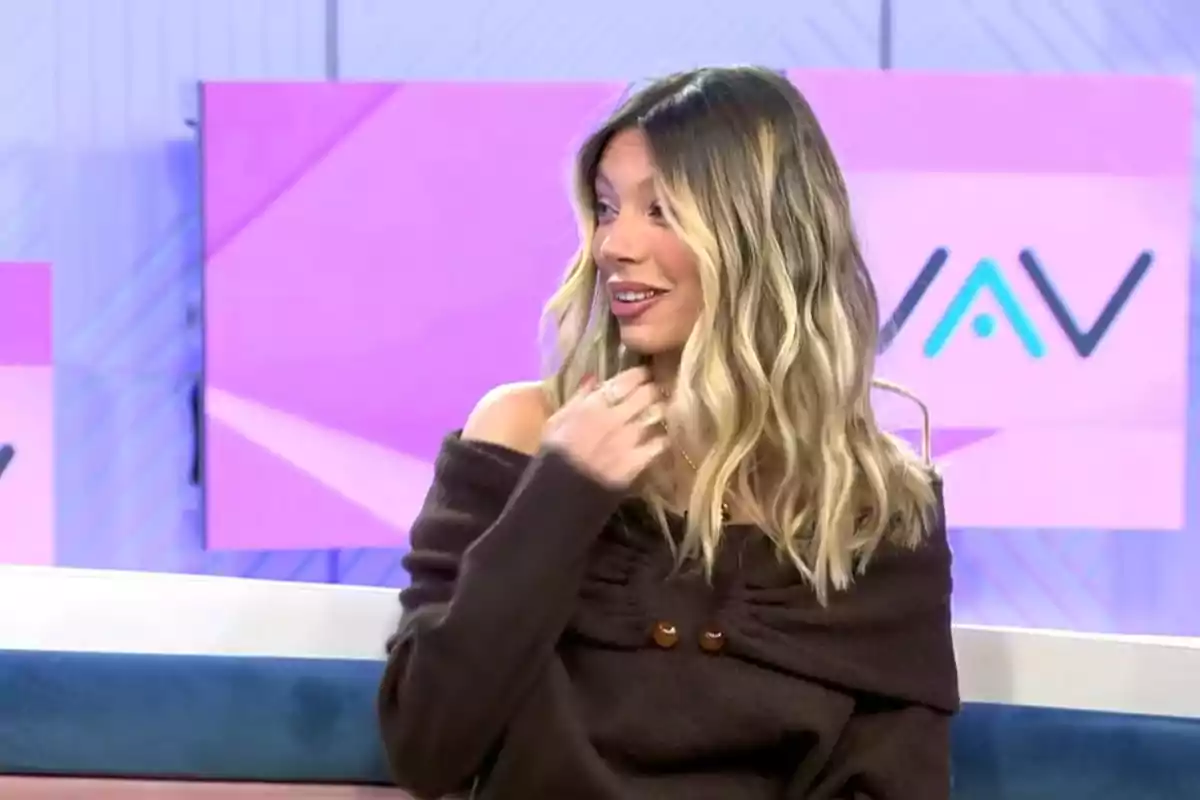 Alejandra rubio con cabello rubio ondulado y suéter marrón oscuro está sentada en un set de televisión con pantallas de fondo en tonos rosados y morados. Captura de 'Vamos a Ver' Alejandra rubio con cabello rubio ondulado y suéter marrón oscuro está sentada en un set de televisión con pantallas de fondo en tonos rosados y morados. Captura de 'Vamos a Ver'