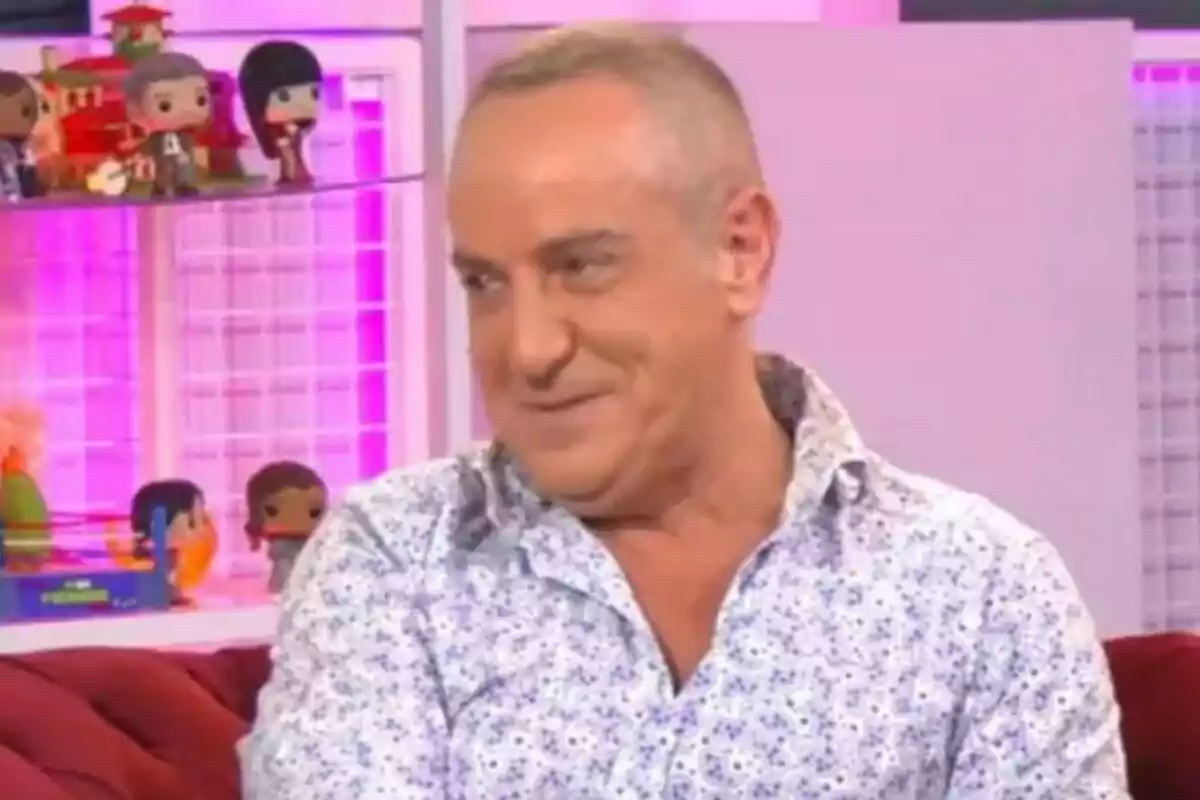 Victor Sandoval amb camisa de flors somriu mentre està assegut en un sofà vermell, amb prestatgeries i figures decoratives al fons a Ni que fuéramos. Victor Sandoval amb camisa de flors somriu mentre està assegut en un sofà vermell, amb prestatgeries i figures decoratives al fons a Ni que fuéramos.
