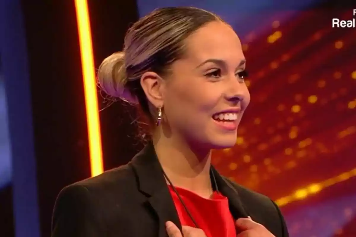 Violeta somrient amb el cabell recollit i vestida amb un blazer negre i brusa vermella en un set il·luminat a Gran Hermano. Violeta somrient amb el cabell recollit i vestida amb un blazer negre i brusa vermella en un set il·luminat a Gran Hermano.
