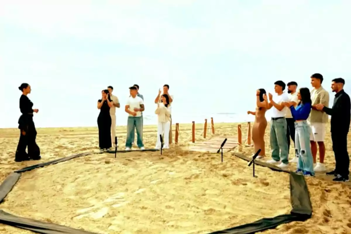 Un grupo de personas está reunido en la playa, algunas aplaudiendo y otras observando, mientras Marina Rivers vestida de negro se encuentra frente a ellos dentro de un área delimitada en la arena en el programa 'Warrior Games'
