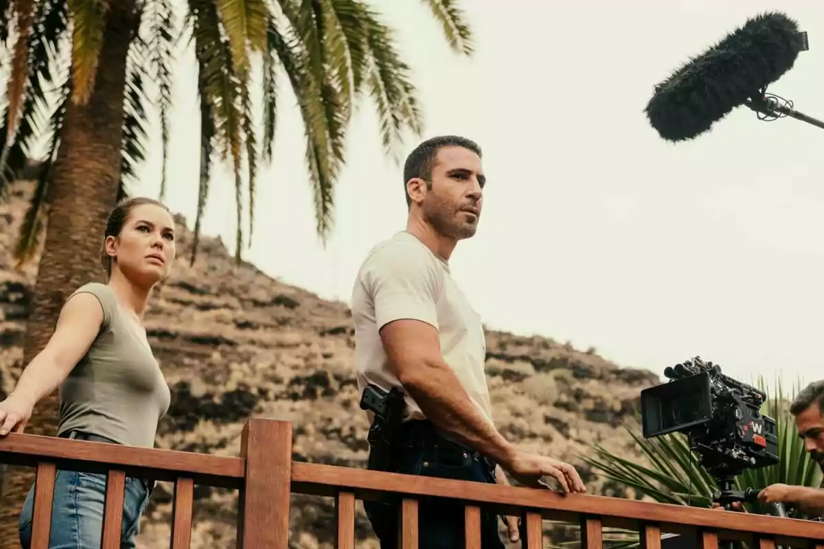 Fotografía de Miguel Ángel Silvestre y Katia Fellin de pie junto a una barandilla de madera, con una cámara de filmación y un micrófono de boom en el fondo, en un entorno al aire libre con palmeras y colinas en el rodaje de Weiss & Morales Fotografía de Miguel Ángel Silvestre y Katia Fellin de pie junto a una barandilla de madera, con una cámara de filmación y un micrófono de boom en el fondo, en un entorno al aire libre con palmeras y colinas en el rodaje de Weiss & Morales