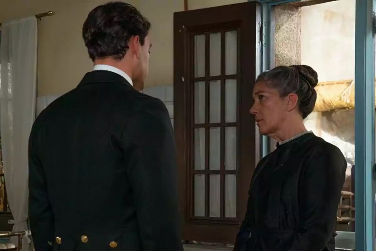Xavi Lock i Marga Martínez estan drets davant d'una porta, mirant-se amb expressions serioses en una habitació il·luminada a La Promesa. Xavi Lock i Marga Martínez estan drets davant d'una porta, mirant-se amb expressions serioses en una habitació il·luminada a La Promesa.