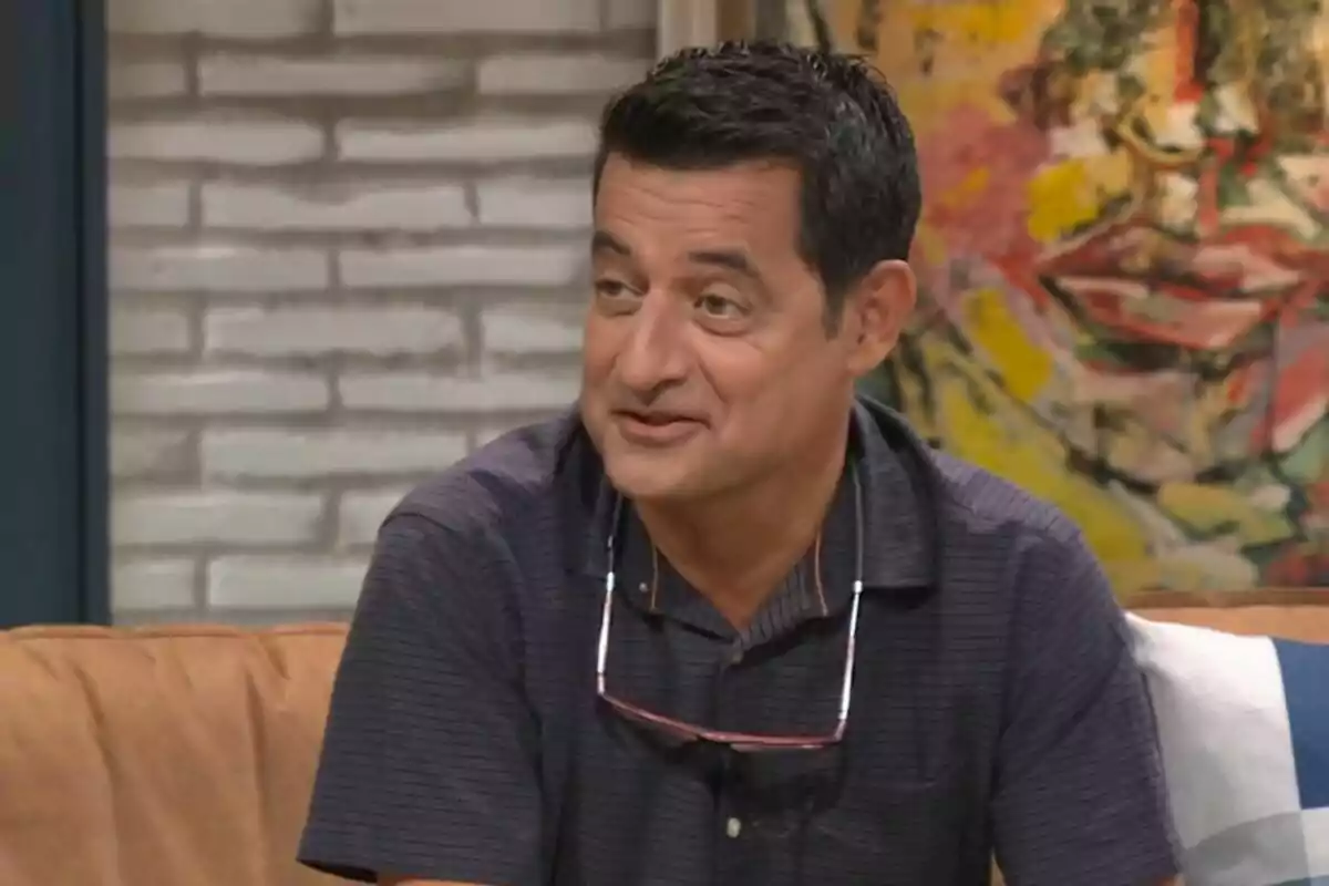 Xavier Bonastre amb camisa fosca i ulleres penjant al coll està assegut en un sofà davant d'una paret de maons i un quadre acolorit.