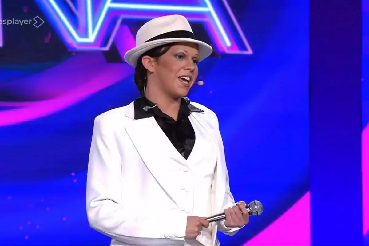 Yenesi vestida amb vestit blanc i barret sostenint un micròfon en un escenari amb fons blau i rosa de Tu Cara Me Suena