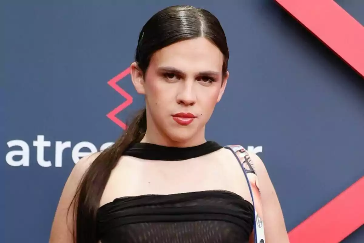 Yenesi con cabello largo y liso, vestida de negro, posa frente a un fondo azul con detalles rojos. Yenesi con cabello largo y liso, vestida de negro, posa frente a un fondo azul con detalles rojos.