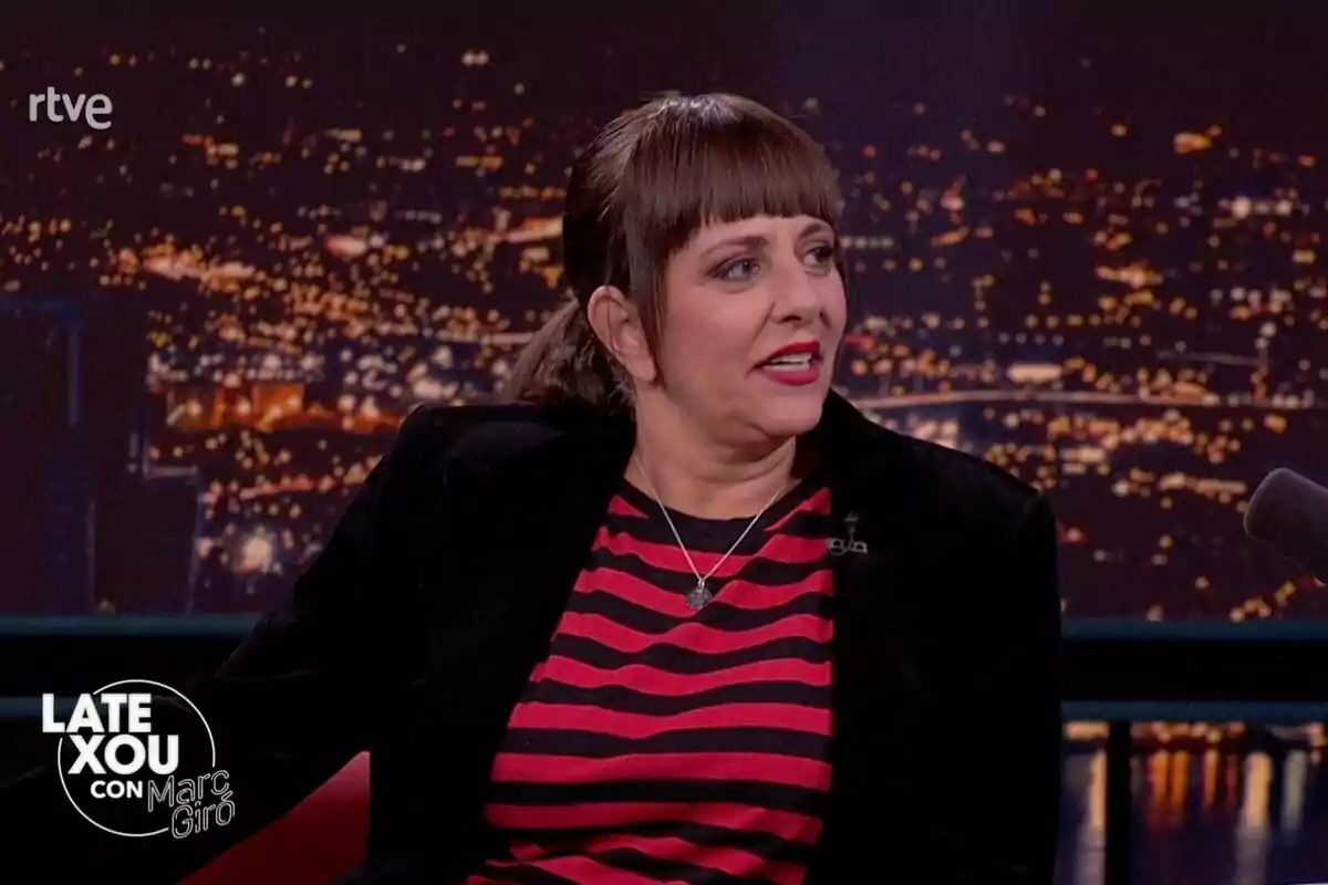 Yolanda Ramos con cabello oscuro y flequillo está sentada en el 'Late Xou' de Marc Giró con un fondo de luces de ciudad, lleva una camiseta de rayas rojas y negras y una chaqueta oscura.