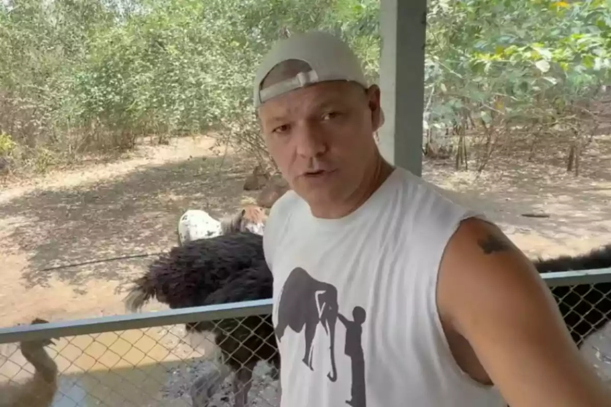 Frank Cuesta con gorra y camiseta sin mangas junto a un avestruz detrás de una cerca en un entorno natural.